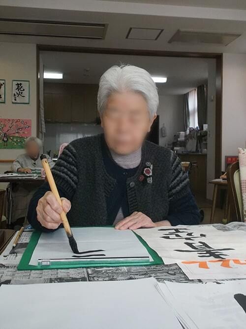 大筆を持ち、見本を見ながら青空という文字を書いている高齢女性。テーブルの上には新聞紙も敷かれている。