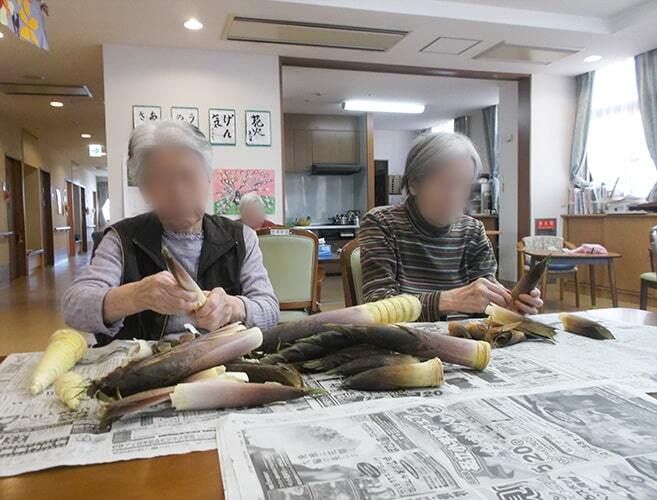 ダイニングテーブルの上には新聞紙が敷かれ、2名の高齢女性たちがタケノコの皮をむいている。