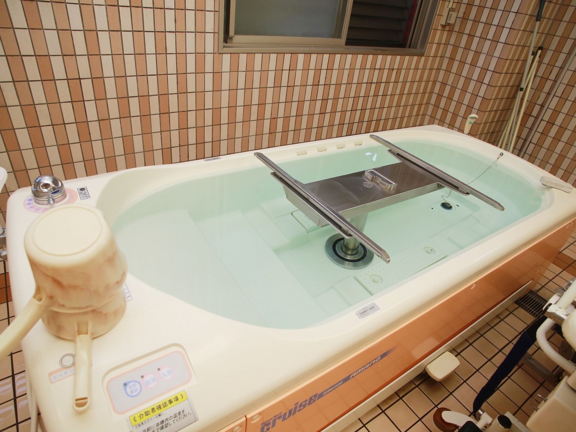 窓のあるタイル張りの浴室に、ステンレス製の昇降器具がついた浴槽が設置してある。手桶が２つ重ねられている。
