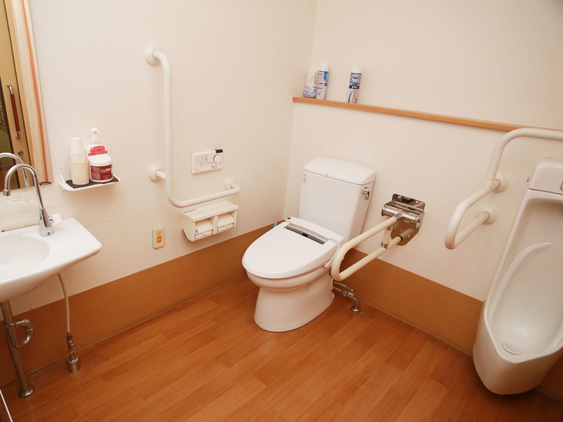 蓋付きの洋式便器と男性用小便器が並べて設置されており、周囲には手すりが施されている。手洗器や鏡が見える。