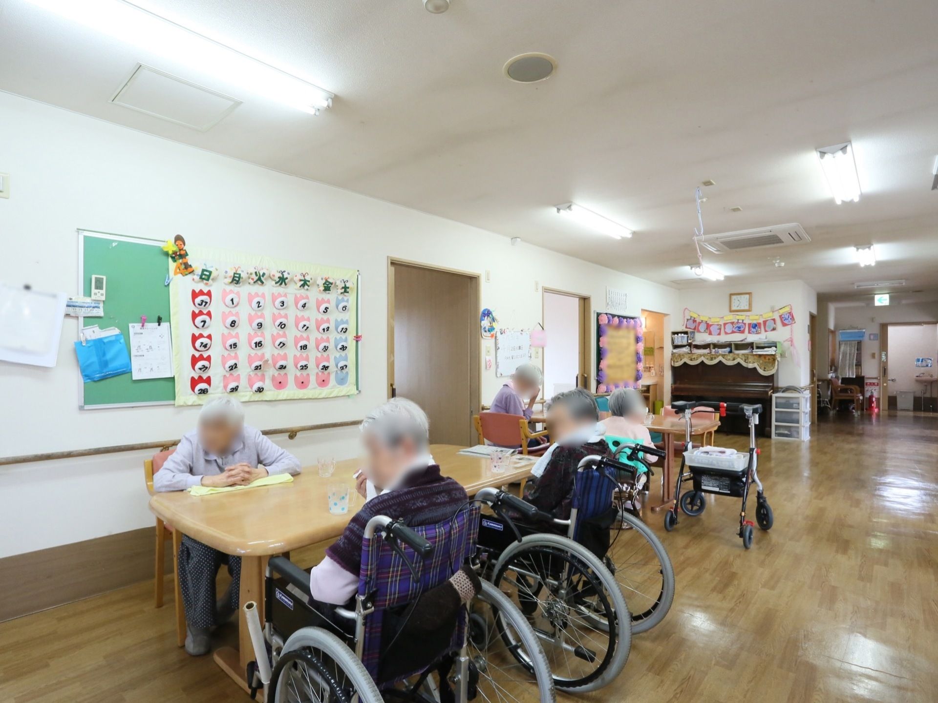 広々とした共有スペースにはテーブルが置かれている。入居者様が椅子に座り、思い思いに過ごしている。壁には手作りのカレンダーが飾られている。