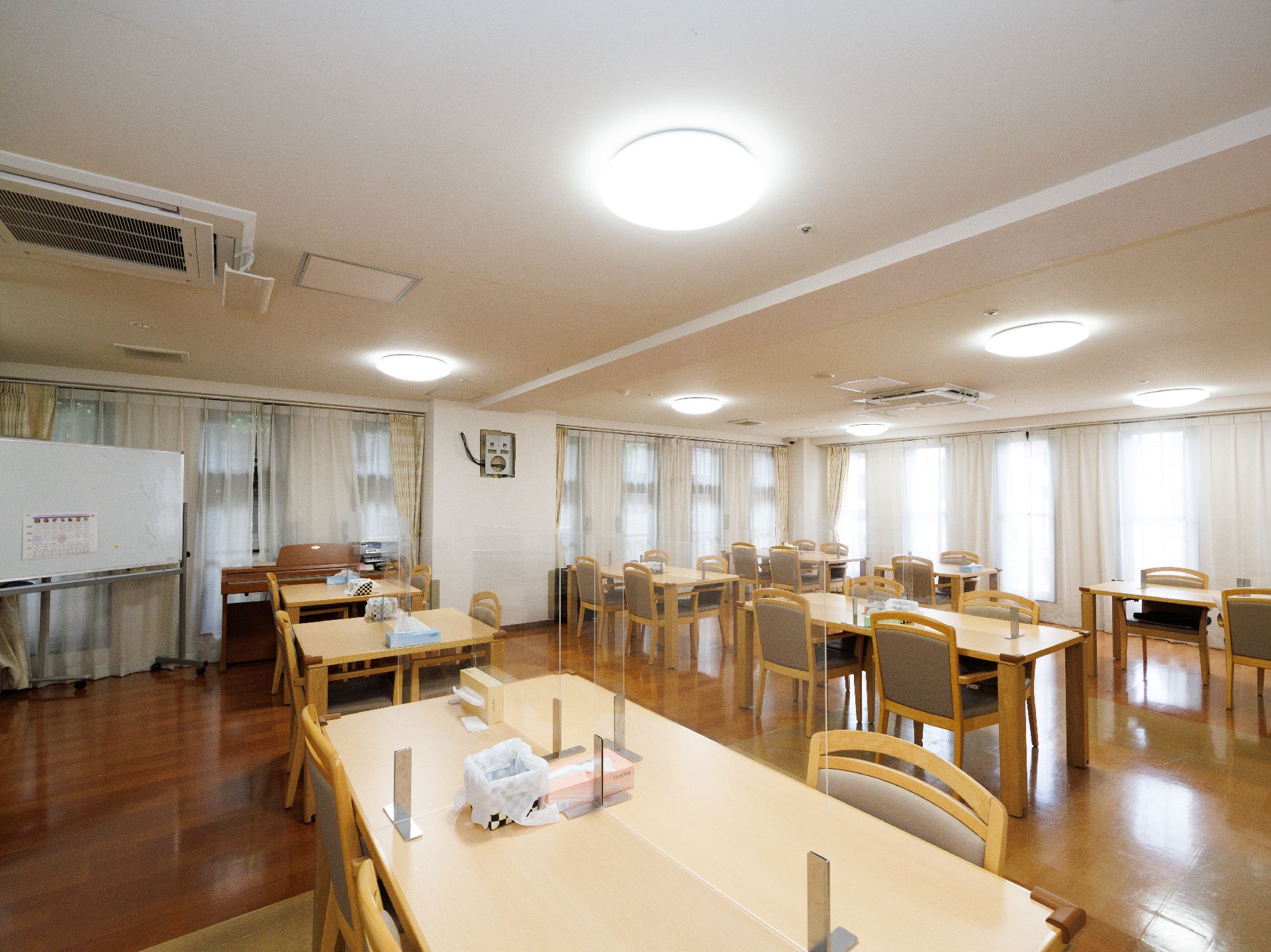 透明な仕切りが付いた木製の机が並んでいる白い壁の広間