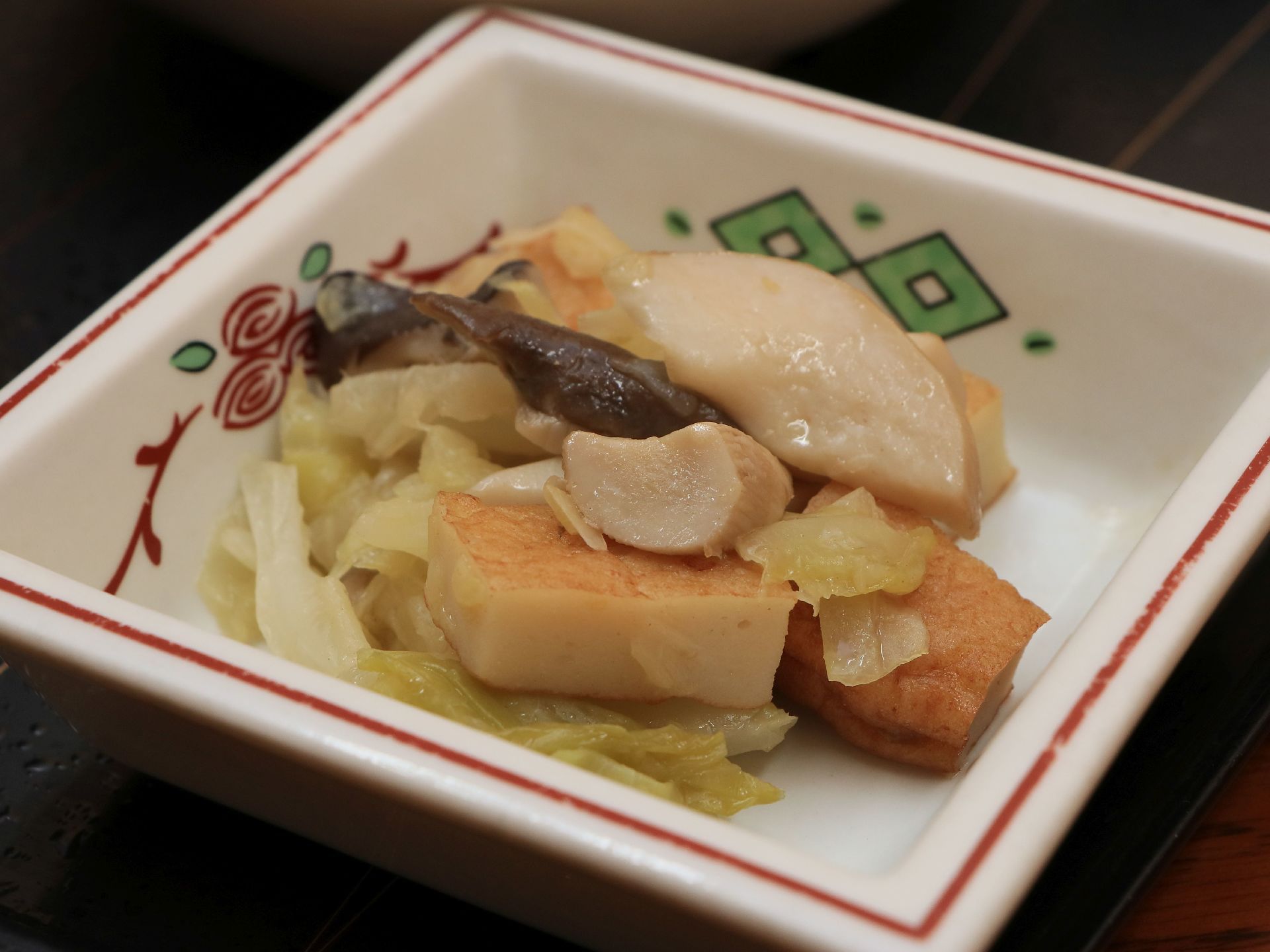 白色で、四角形の小鉢に煮物が盛りつけられている。食べきりやすい量である。食材は葉物や練り物などが使われている。