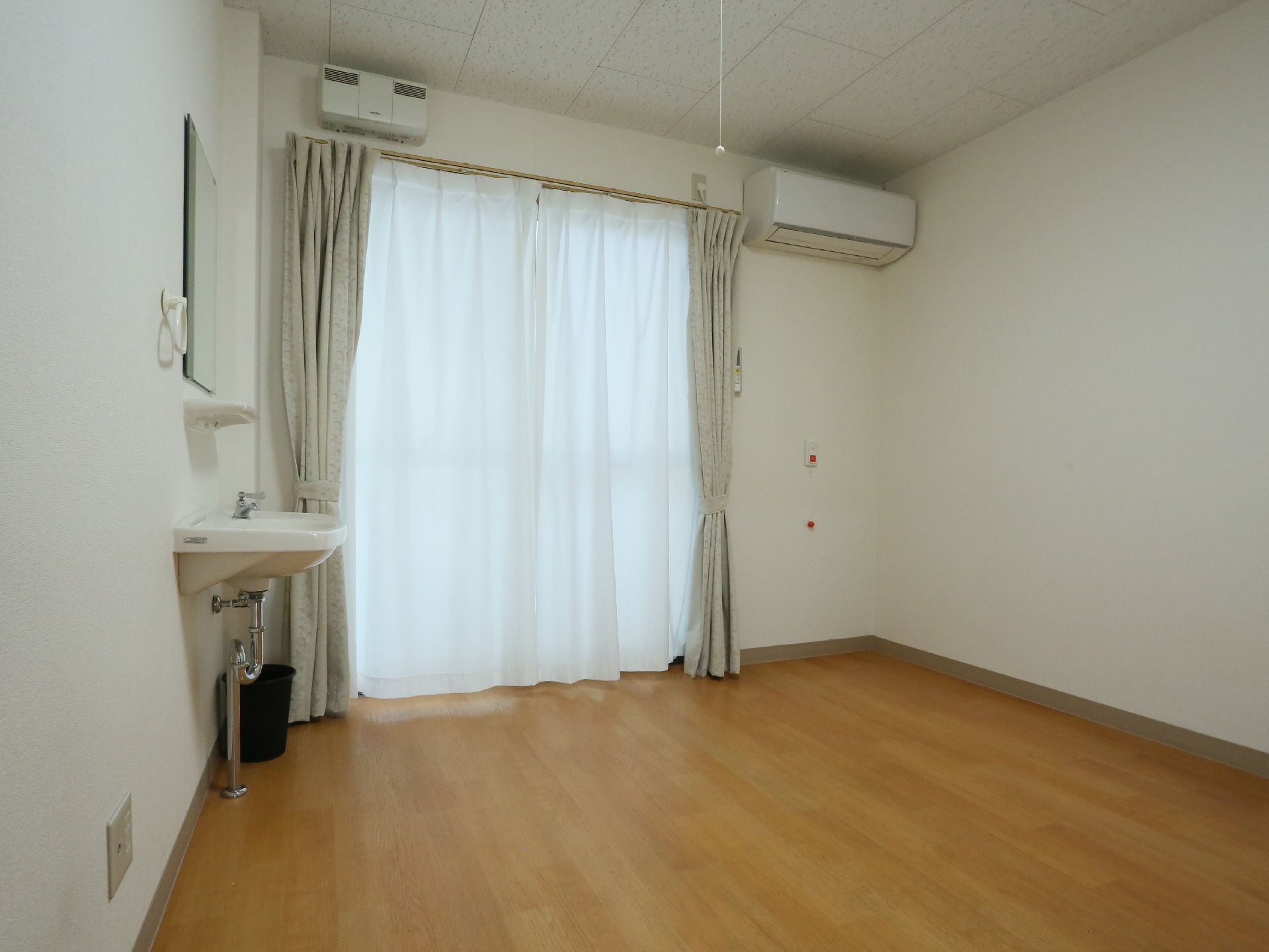 居室には掃き出し窓がある。エアコン、ナースコールボタン、洗面台が設置されている。洗面台の前の壁には鏡がある。