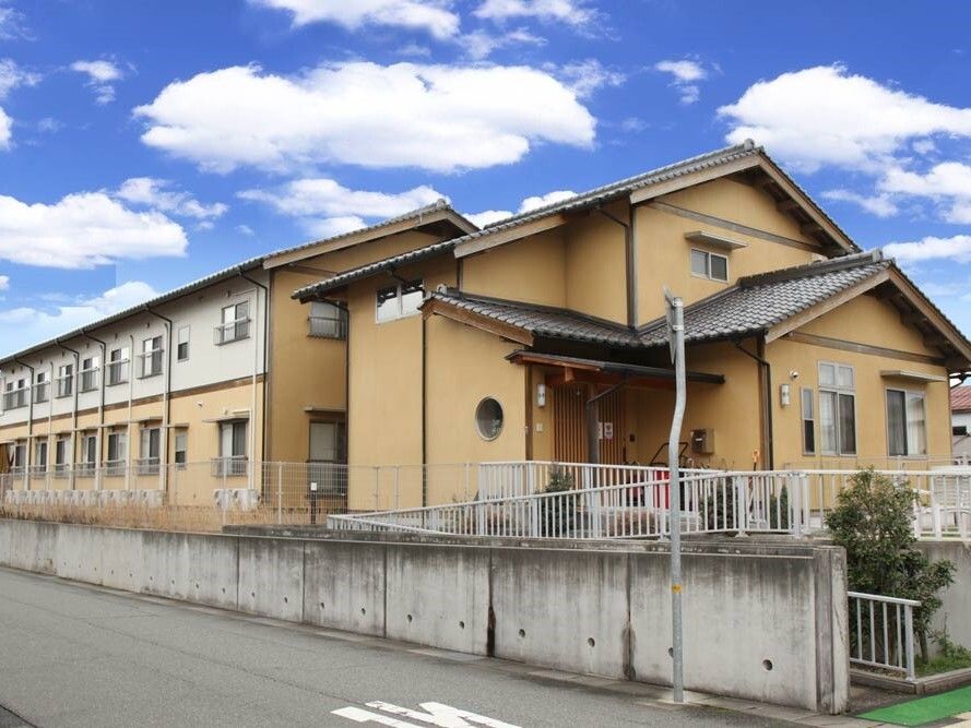 三角屋根が付いた２階建ての建物は日当たり良好で建物の周りにはフェンスがあり、敷地内には草木が植えられている。