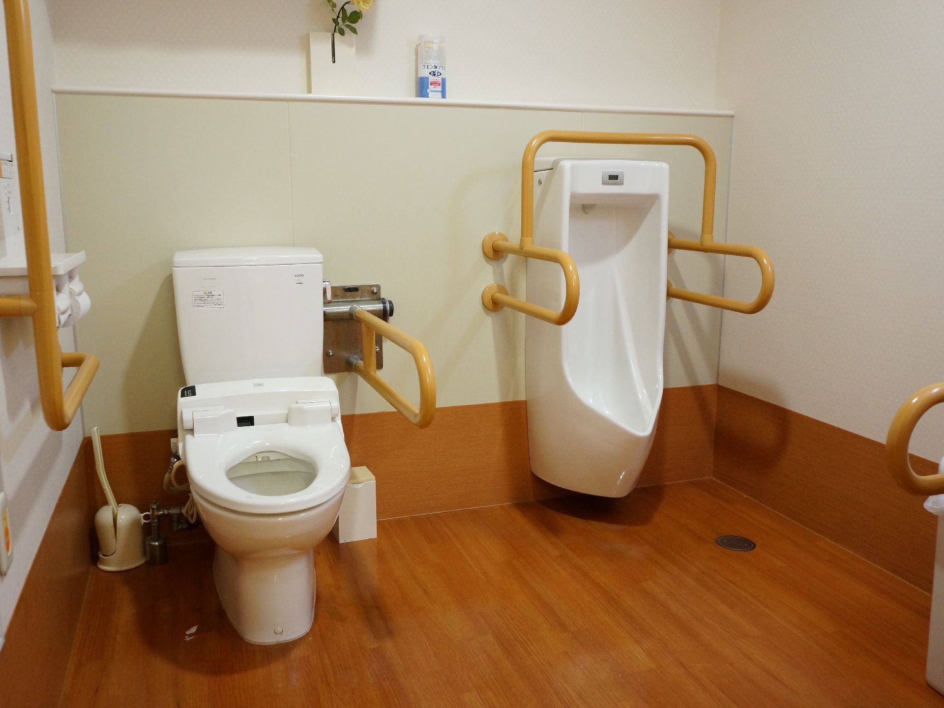 白い壁と木目調のフロアがスッキリとして清潔感があるトイレ。洋式便器と小便器が設置された広々とした空間になっている。