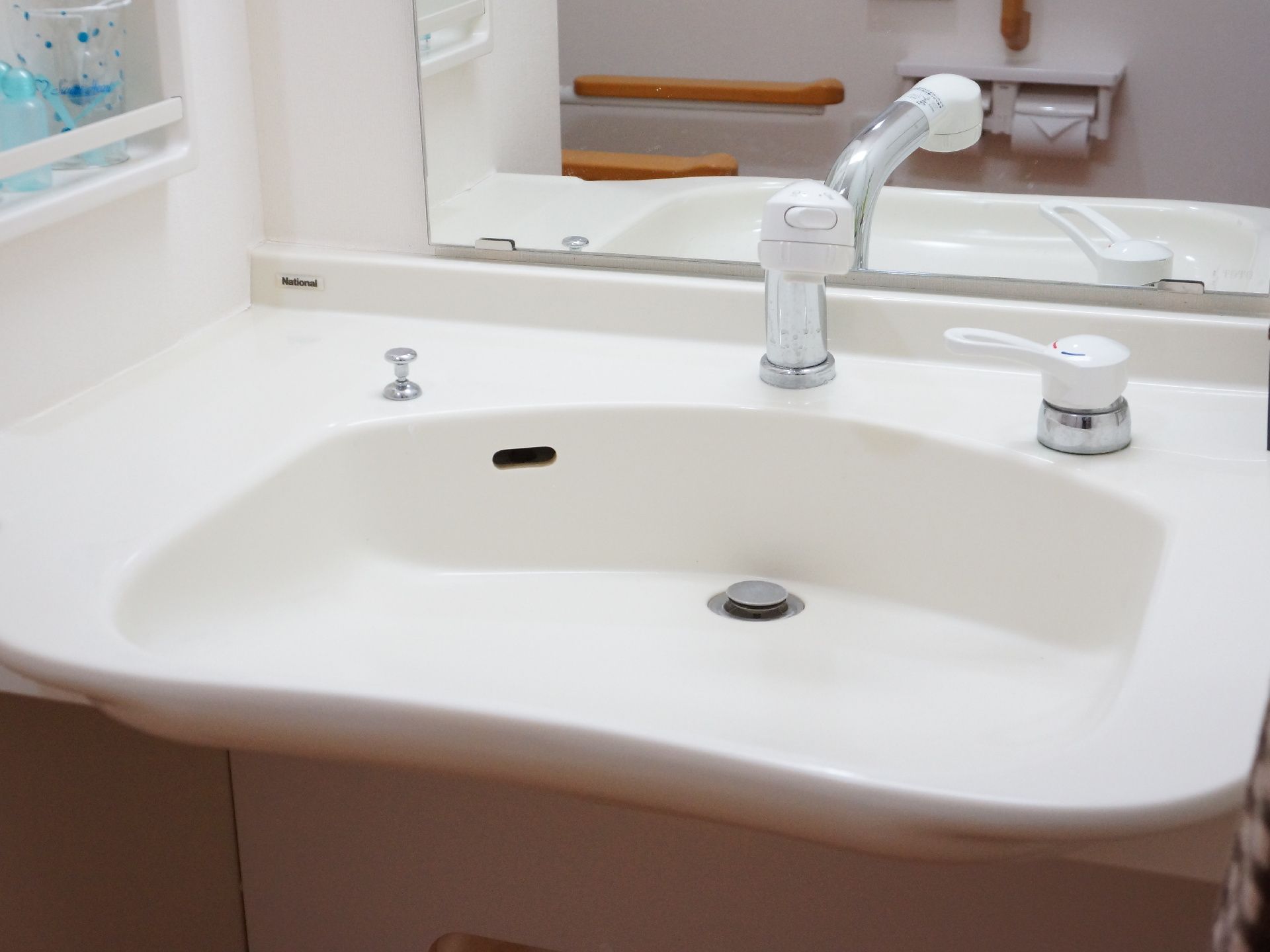 洗面台は白く、前方に鏡が取り付けられている。鏡には手すりとトイレットペーパーフォルダーが写っている。