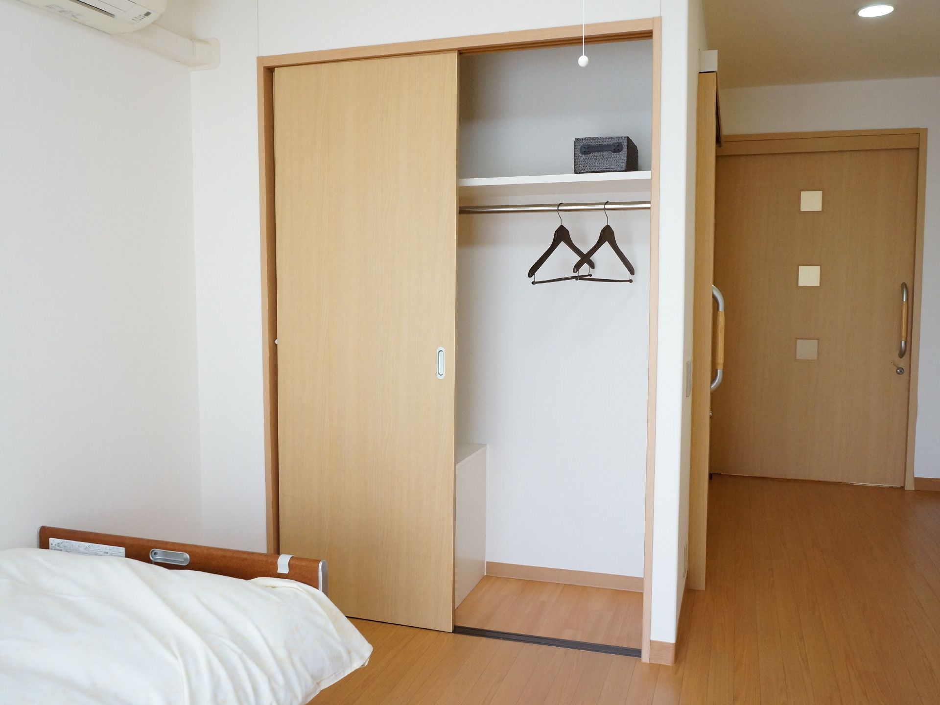 居室には茶色い枠のベッドが置かれている。足元側にはクローゼットが設置されている。中にはパイプハンガーが取り付けられている。
