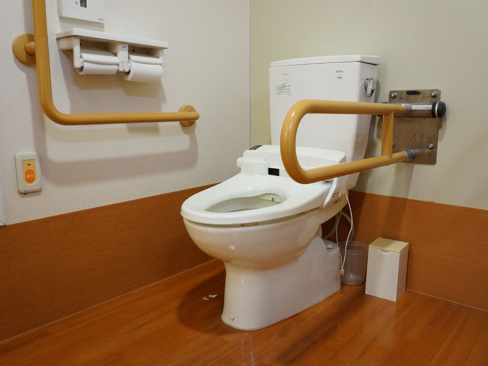 白い壁と茶系のフロアに置かれている洋式便器。便器の両脇には手すりが設置され、壁には緊急の場合の通報装置が取り付けられている。