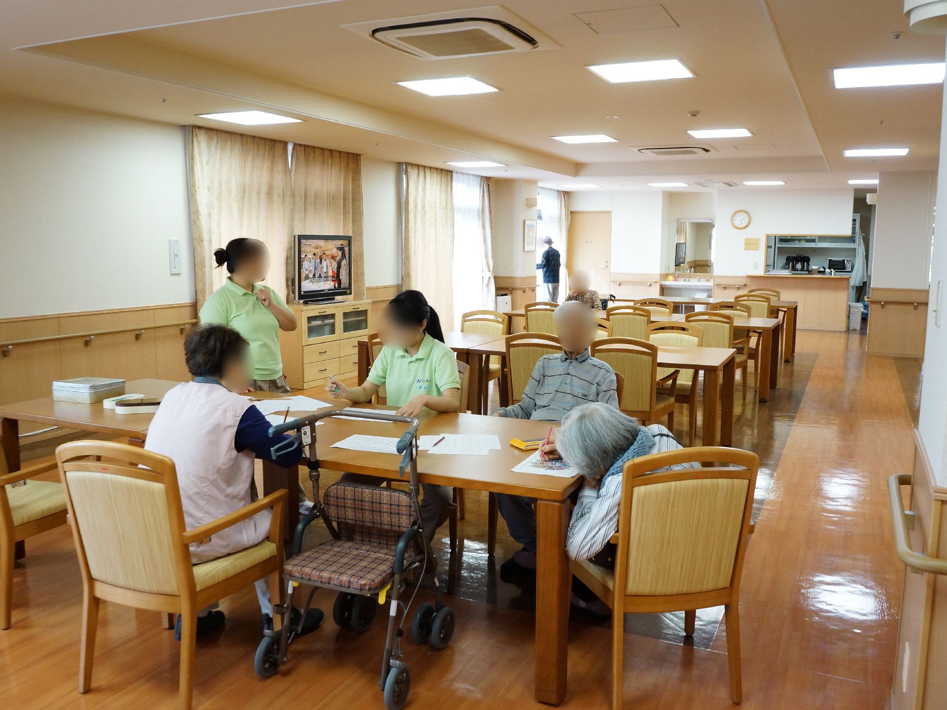 リビングルームには茶色いテーブルと椅子が並べられていて、椅子にはスタッフと入居者様が座って作業をしている。壁には手すりが設置されている。
