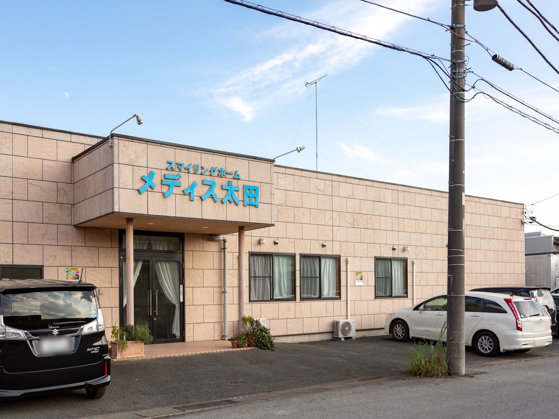 外壁はベージュのタイル貼りで、建物の前に車が停まっている。前方には道路があり、電信柱が１本立っている。