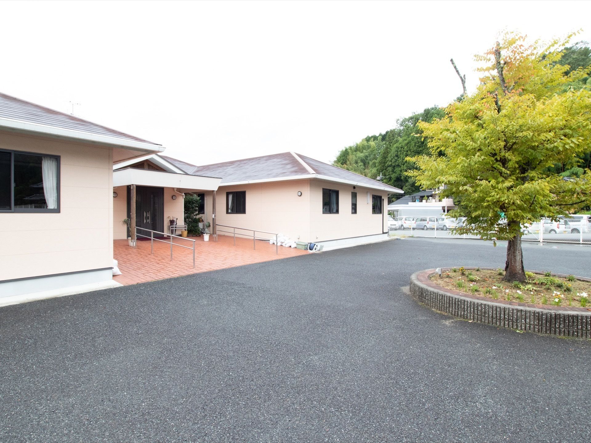 施設のエントランス前は、コンクリート舗装された敷地がゆったりと広がっている。中央には大きな木が1本、円形の花壇に植えられている。