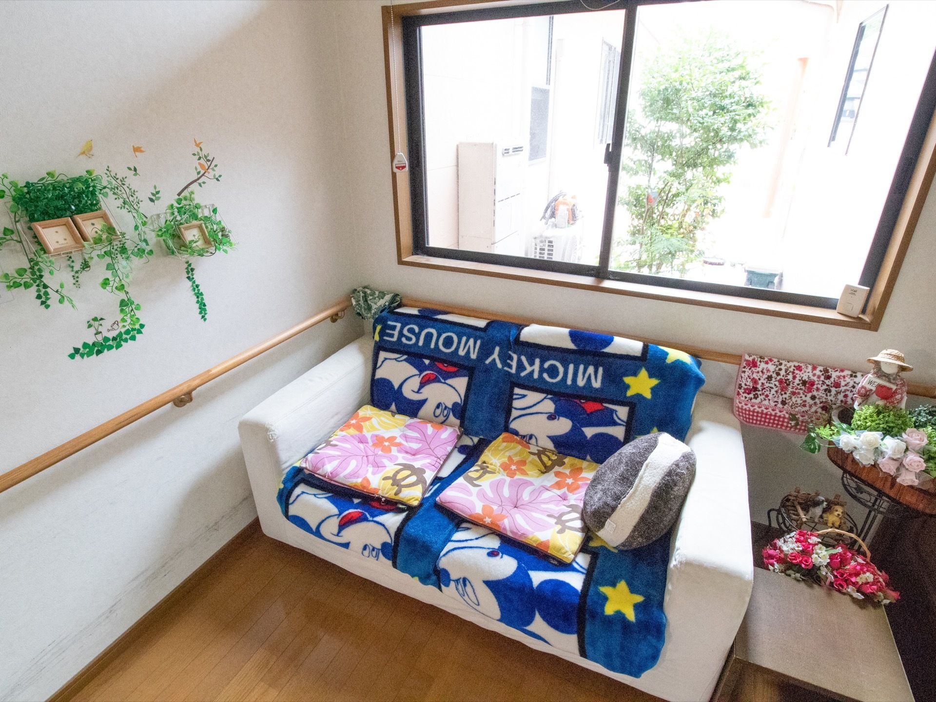 窓際にソファが置かれている。ソファの横や近くの壁には花や緑、お人形が飾られている。手すりが壁に設置されている。