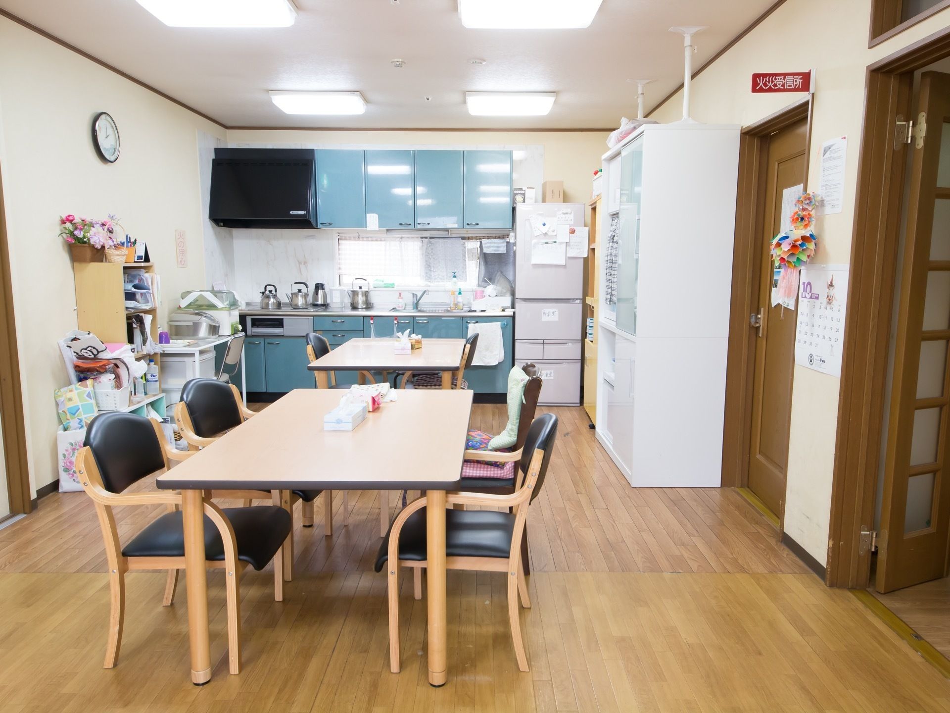 キッチンが設置されており、冷蔵庫や収納棚が備えられている。壁にはカレンダーが掛けられており、アットホームな雰囲気の食堂になっている。