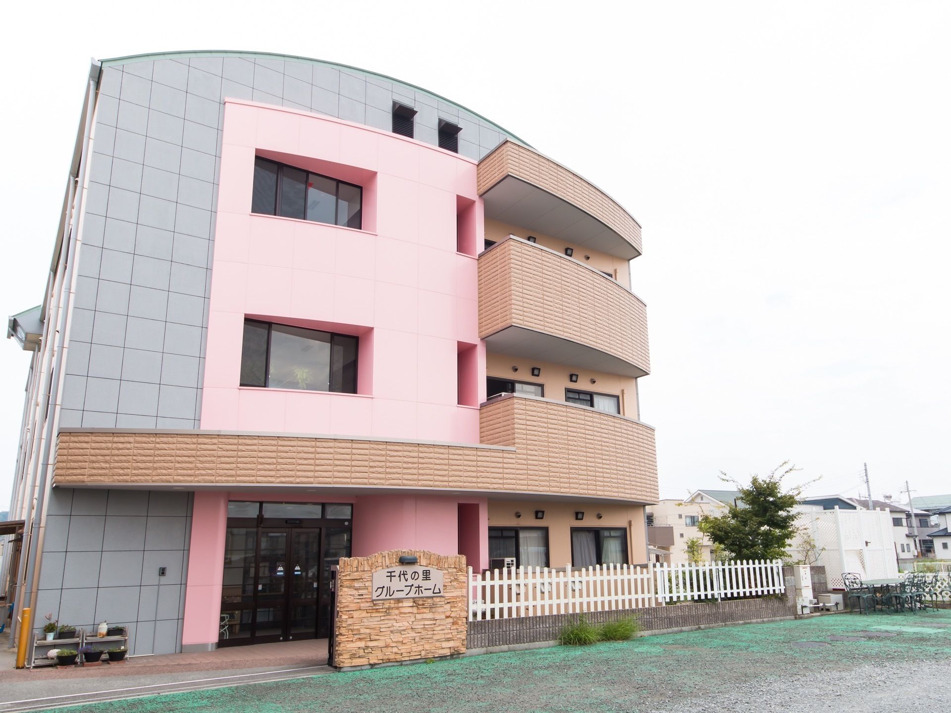 優しいグレーやピンク、茶色を使用した外壁の3階建ての施設である。周辺は閑静な住宅地で、高い建物などはない。