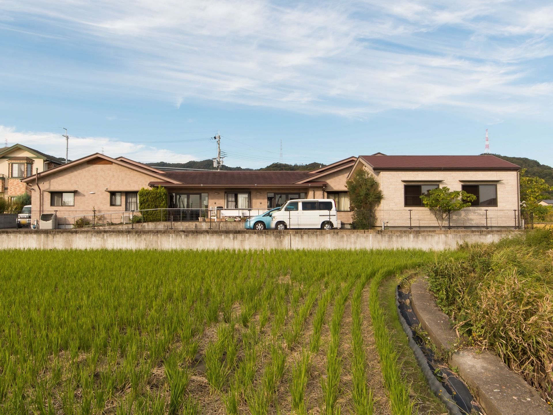 周囲を田んぼや木々、住宅などに囲まれた平屋建ての大きな建物がある。建物の周囲には駐車場があり、敷地の周囲にはフェンスがあるのが見える。