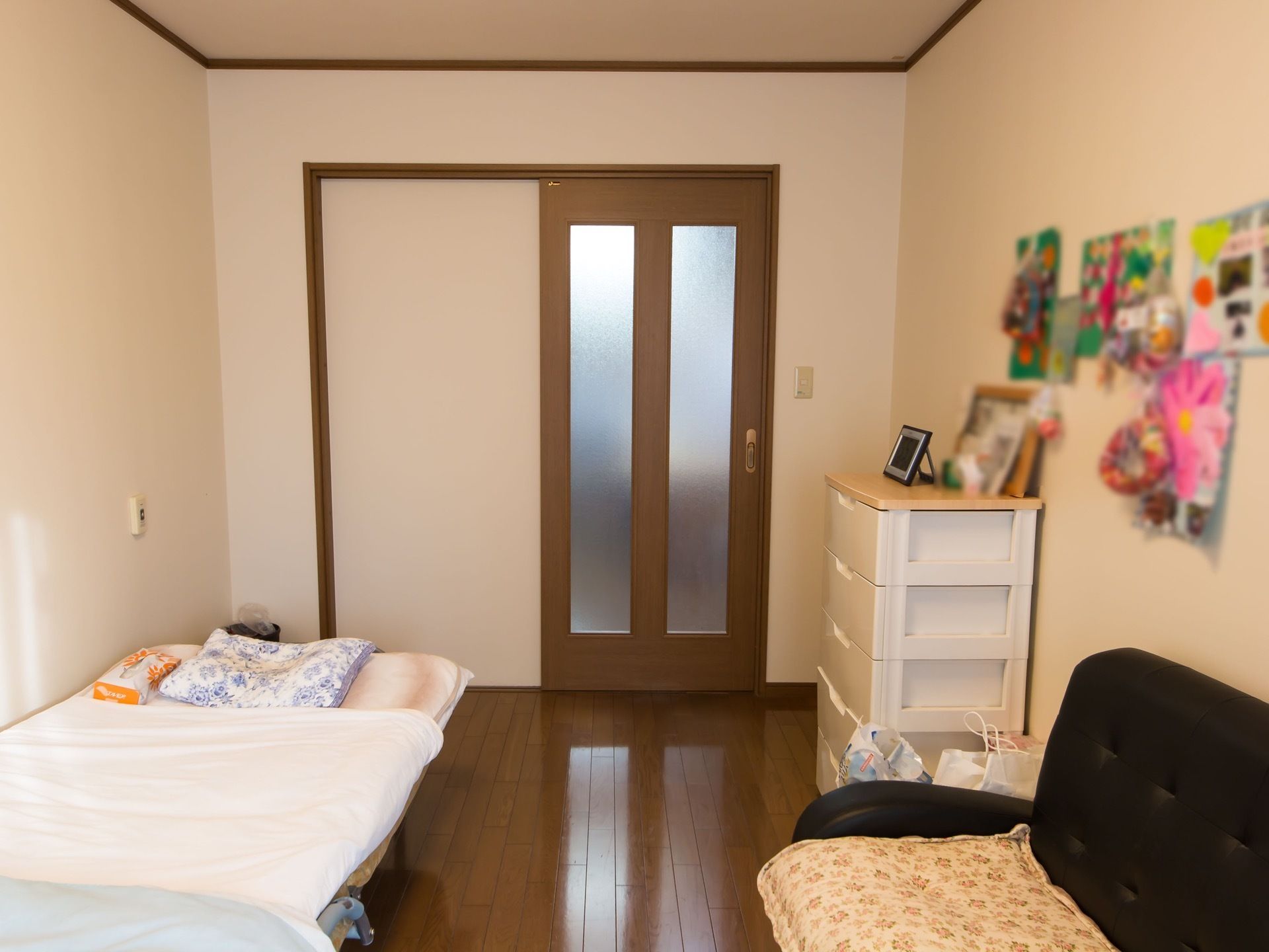 濃い茶色のフローリングの床の居室。入口の扉は引き戸になっている。室内にはベッドと収納家具、ソファが置かれている。