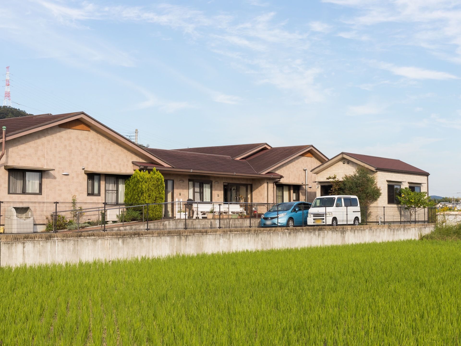 緑豊かな田畑や山などに囲まれた平屋建てのホーム。ホームの前には手すり付きのスロープがあり、駐車場に車が２台停められている。