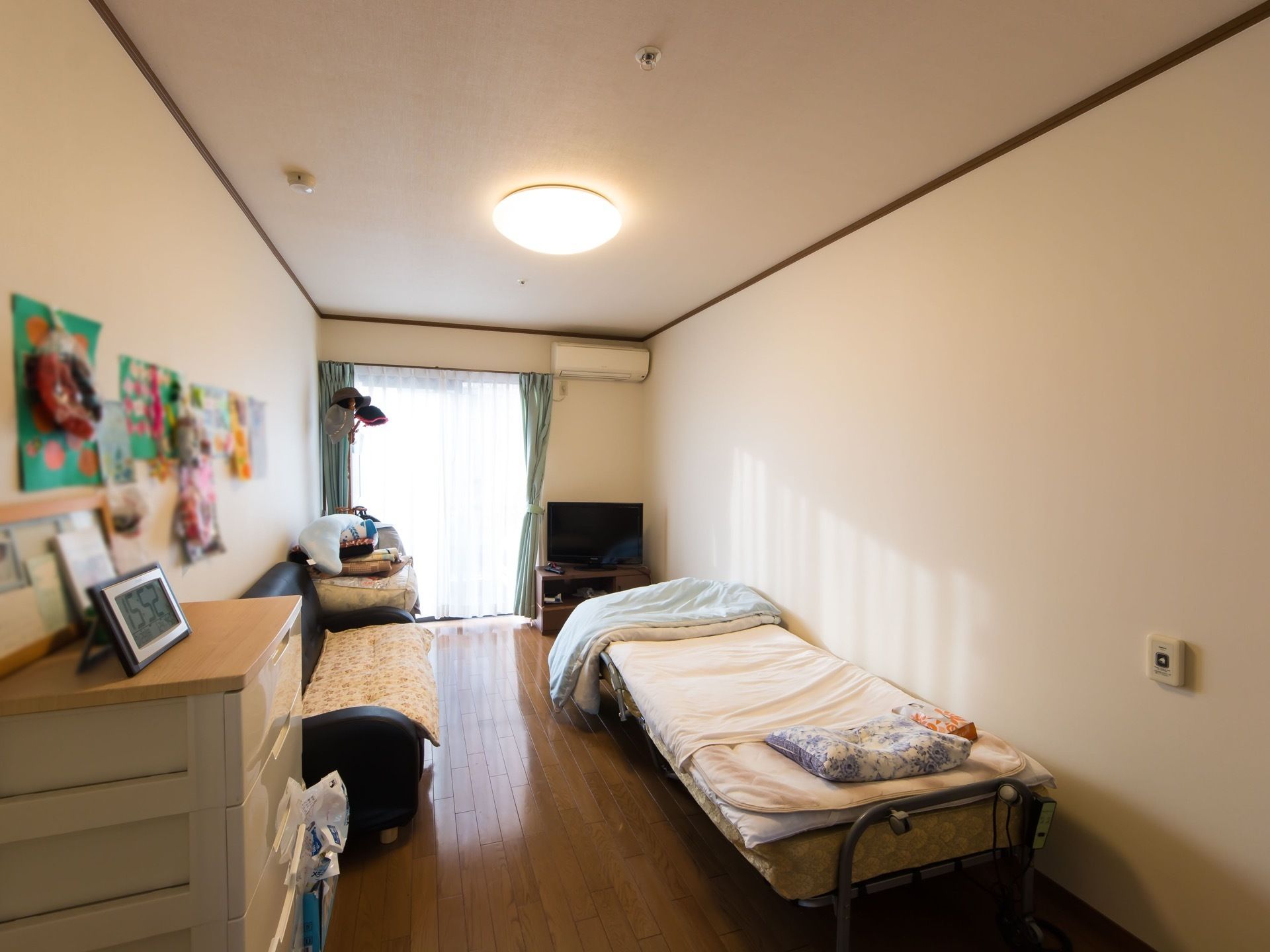 広い居室にベッドと収納、ソファ、テレビとテレビ台などが置かれている。壁にはエアコンも備え付けられている。