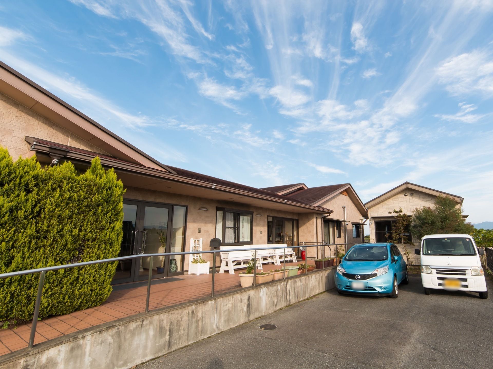 集合住宅風の外観の大きな平屋建ての建物があり、玄関前にはテラスがあるのが見える。テラスの横には駐車場も設けられている。
