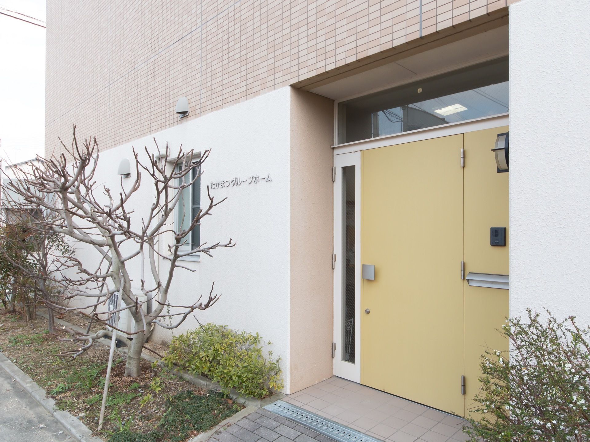 クリーム色の大きな扉で段差はない。玄関先には背丈の低い草木が植えられている。壁に施設名が書かれている。