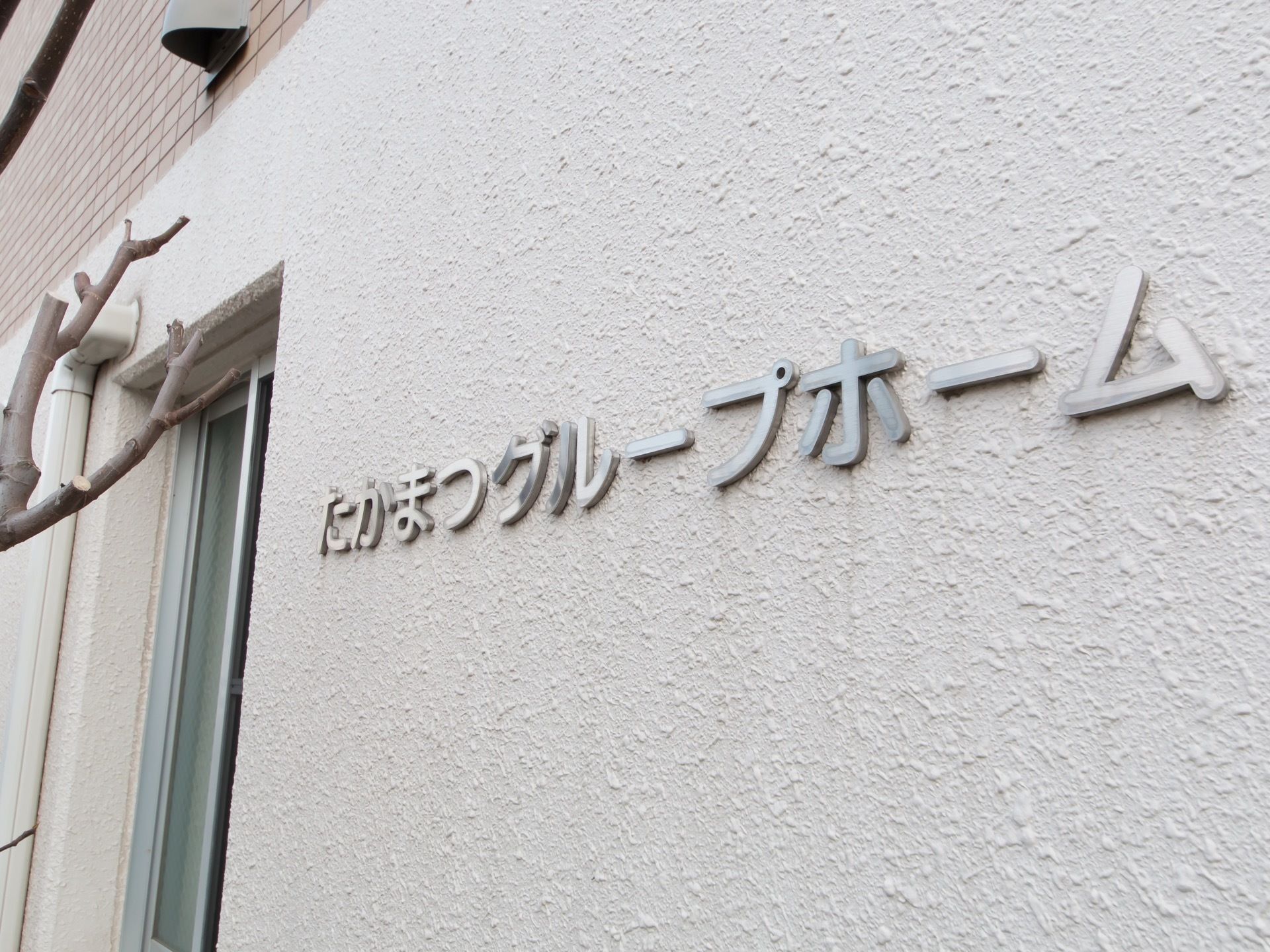 白い壁に銀色の文字で、施設名が取りつけられている。施設周囲には木が植えられており、剪定された枝が見える。