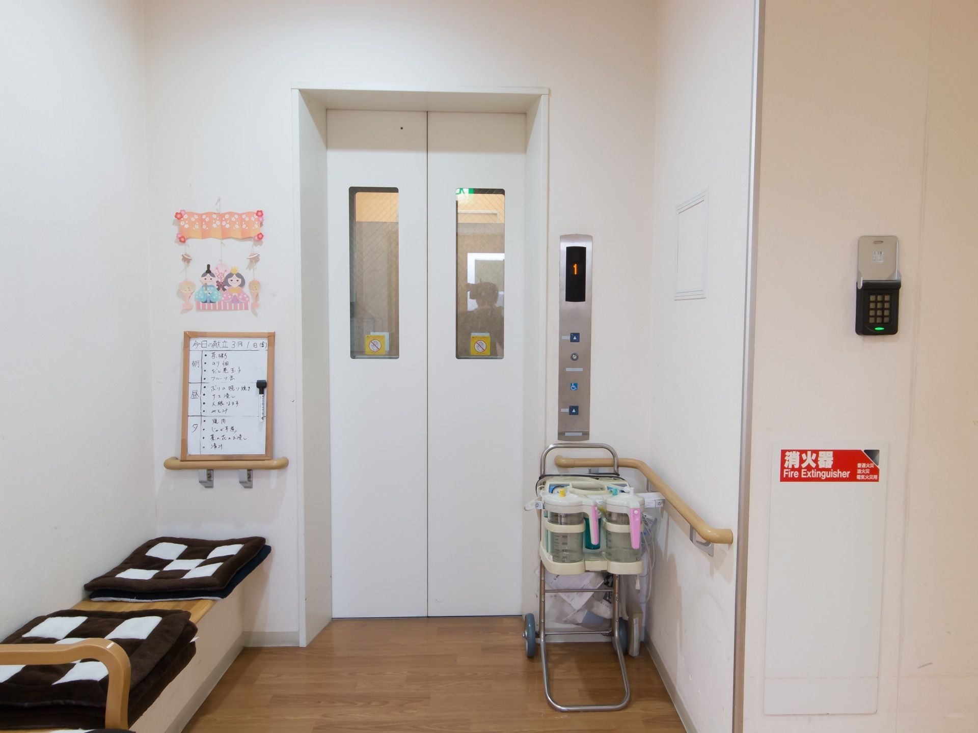 壁と同じ白色のエレベーターである。壁にはベンチがあり、その横に献立表が立て掛けられている。ひな祭りのクラフトが飾られている。