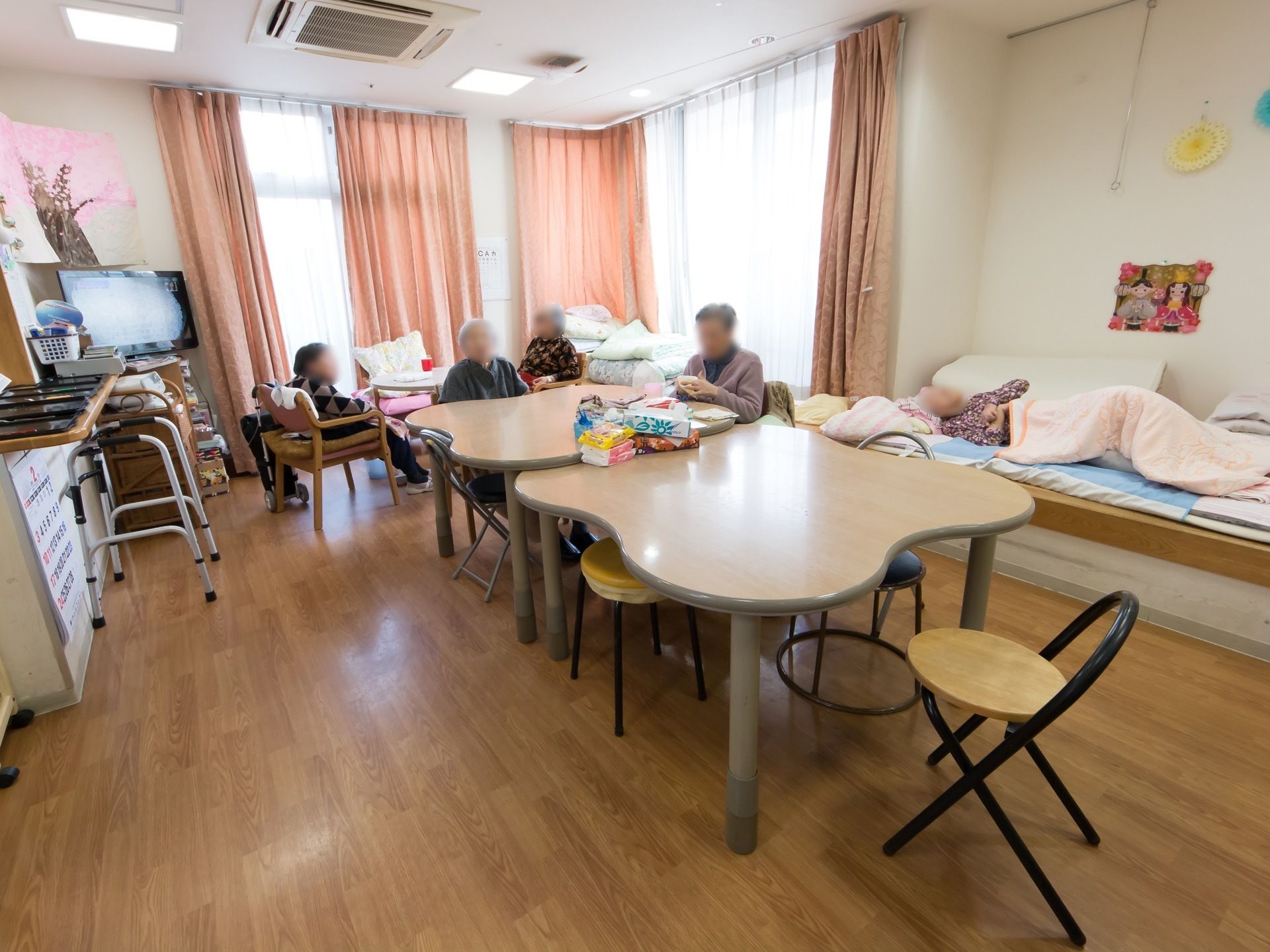 変形テーブルが中央に置かれ、テーブルを囲むように入居者様が座っている様子である。大きな窓があり、ピンクのカーテンがかけられている。