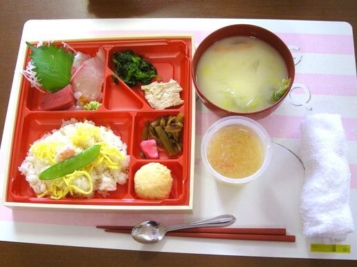 赤い弁当箱にはちらし寿司や刺身、漬物などが詰められ、その横にはみそ汁やおしぼりが置かれ、箸とスプーンが添えられている。