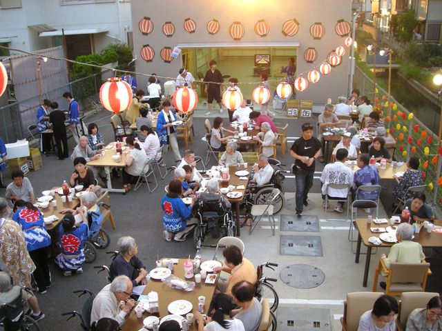 提灯がたくさん飾られた屋外のスペースにはテーブルが並べられ、たくさんの人が食事をしている。スタッフはハッピ姿。