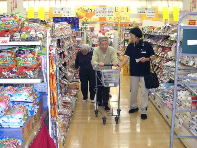 スーパーマーケットのお菓子売り場で買い物を楽しむ女性入居者２名と付き添いの女性スタッフ。