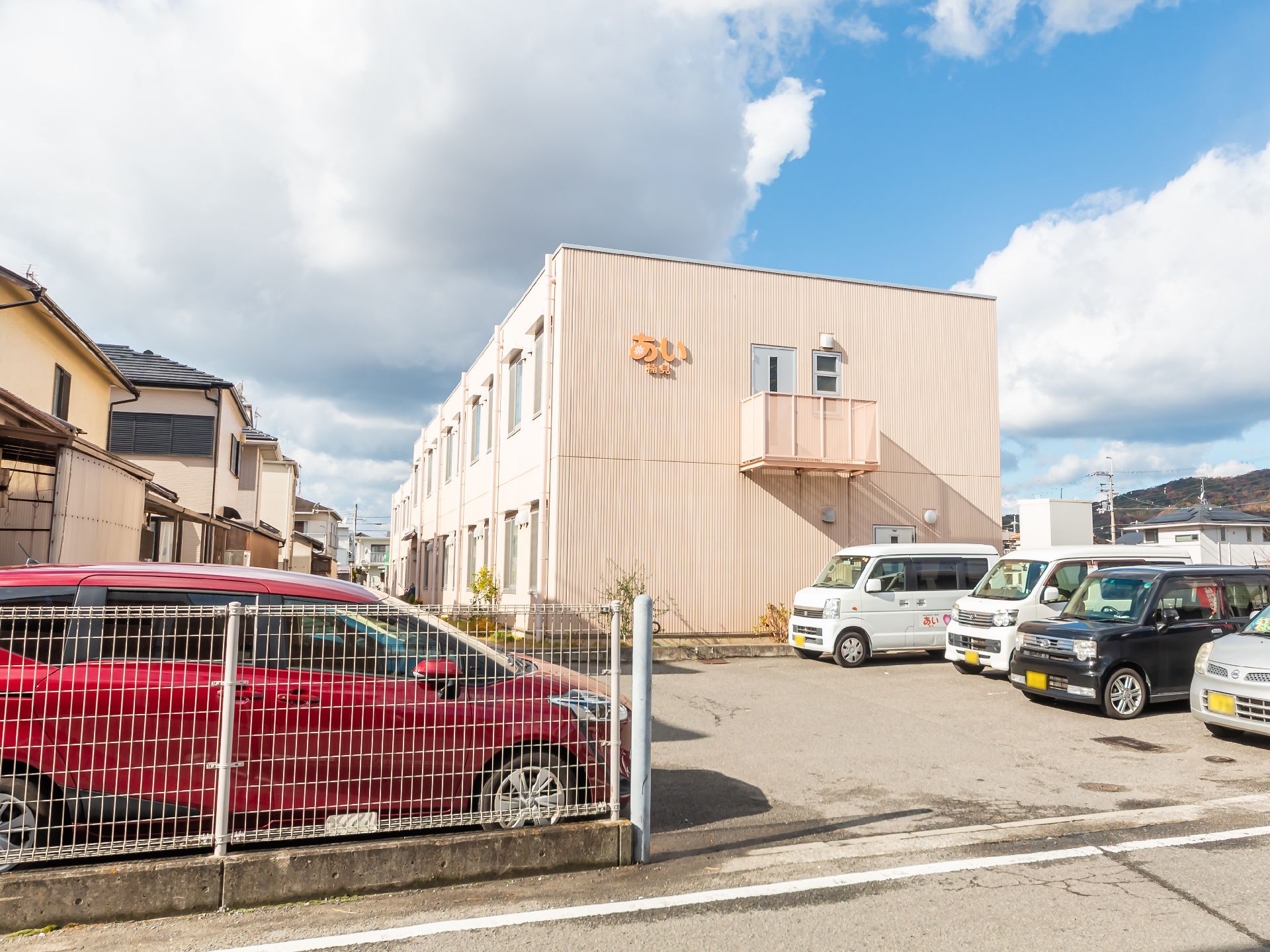 ベージュの2階建ての建物の周りにはベンチが置かれ、複数名の人が座っている。敷地内には広い駐車場も完備されている。