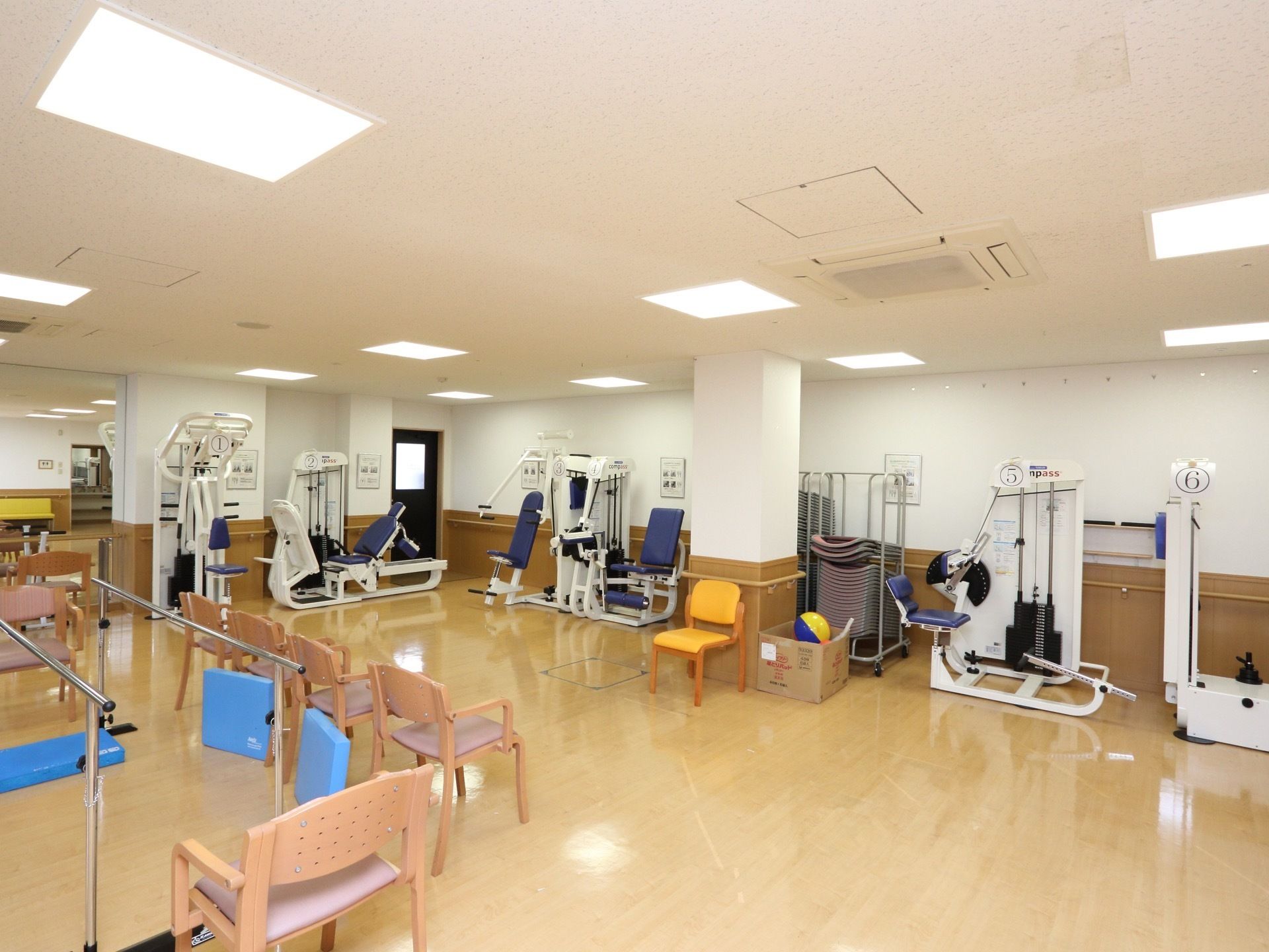 施設内に広いスペースのリハビリ室がある。照明で明るい部屋の中に、多くのリハビリ器具や椅子が備えつけられている。
