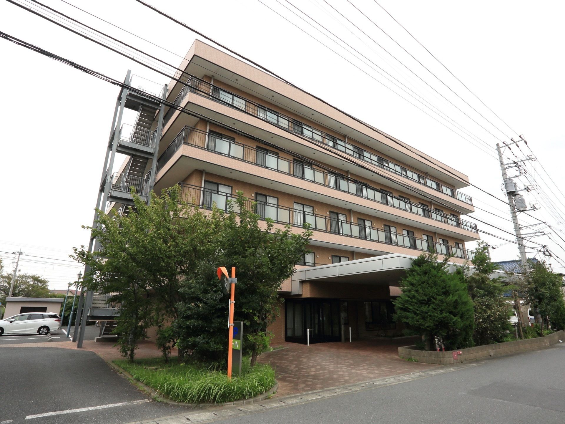 落ち着いた色調の５階建ての施設である。玄関前には緑豊かな植栽があり、側面部には避難階段や駐車場がある。