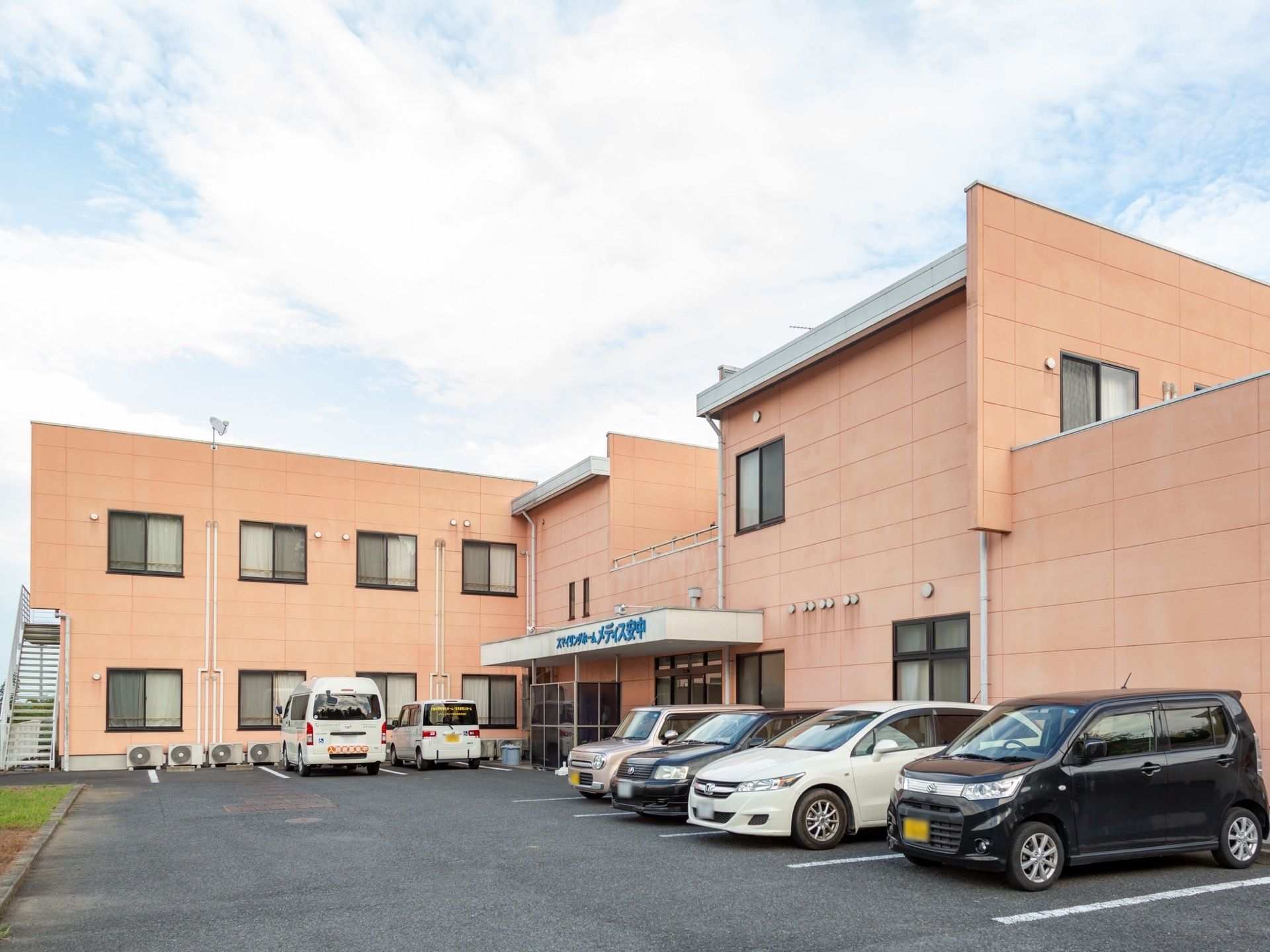施設は外壁の色が暖色系になっている。建物の前に駐車場が完備されており、送迎用のワゴンなど、数台の車が停まっている。