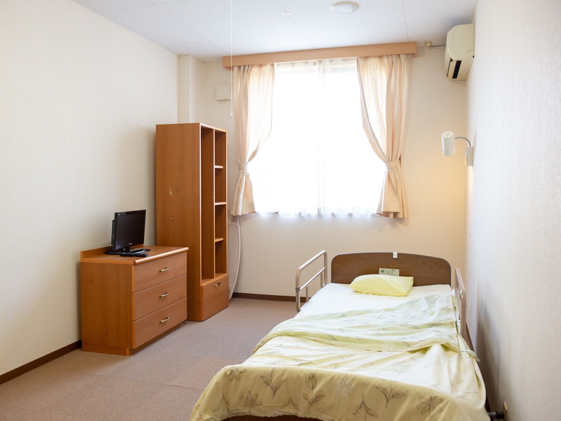 居室には介護ベッドやタンスのほか、服を掛けたり本をおいたりできる棚などが置かれている。床には絨毯マットが敷かれている。