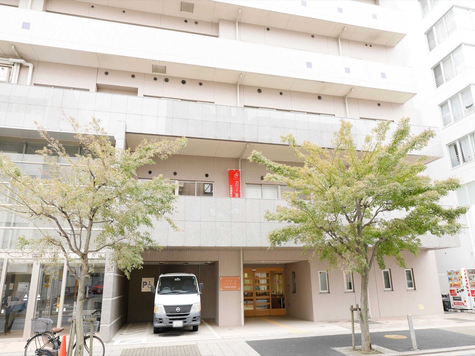マンションのような大きく白い建造物である。玄関の横に大型車が駐車されている。木が植えられており、そこに自転車が置かれている。