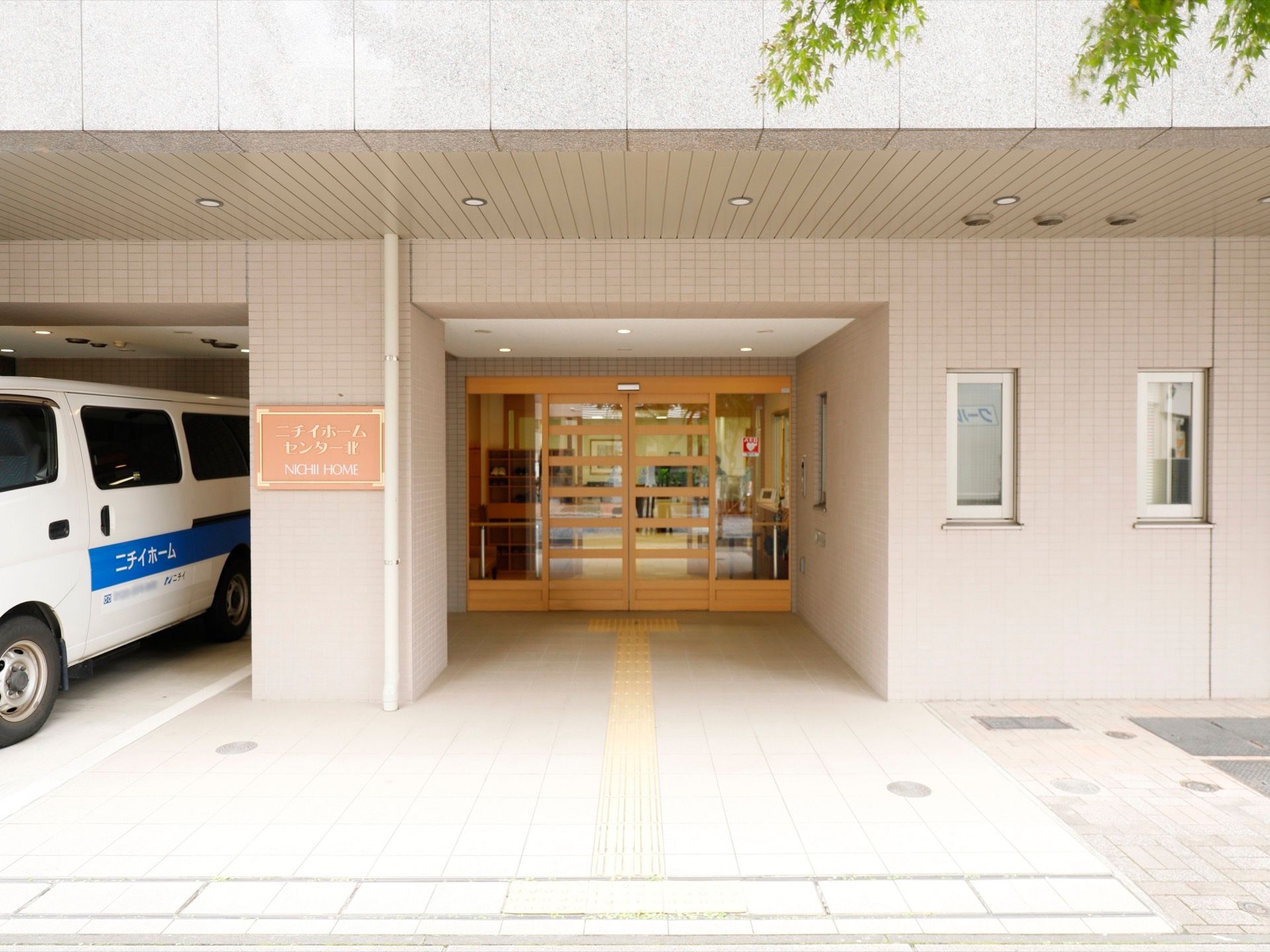 間口の広い黄褐色の自動扉である。その横に施設専用の駐車スペースが用意されている。施設名が書かれた看板は薄いピンク色である。
