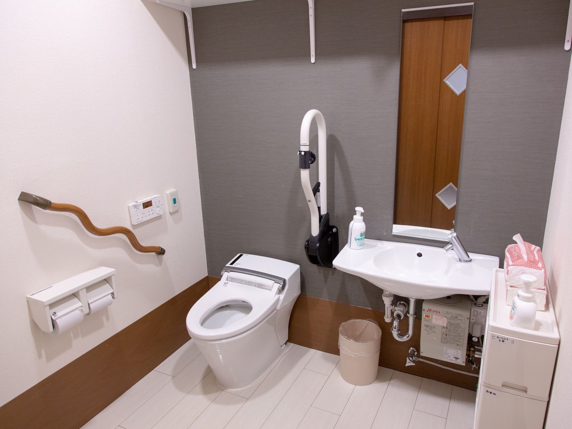 タンクレスの洋式便器と洗面台が並んで壁に設置されている。便器の両側に手すりが付いており、ゴミ箱と収納ボックスが置かれている。