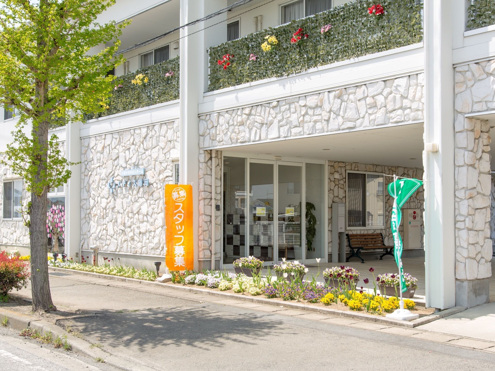 ２階のベランダの柵にはフェイクの植物の目隠しが貼られている。花が植えられた花壇にスタッフ募集のオレンジ色の旗が立っている。