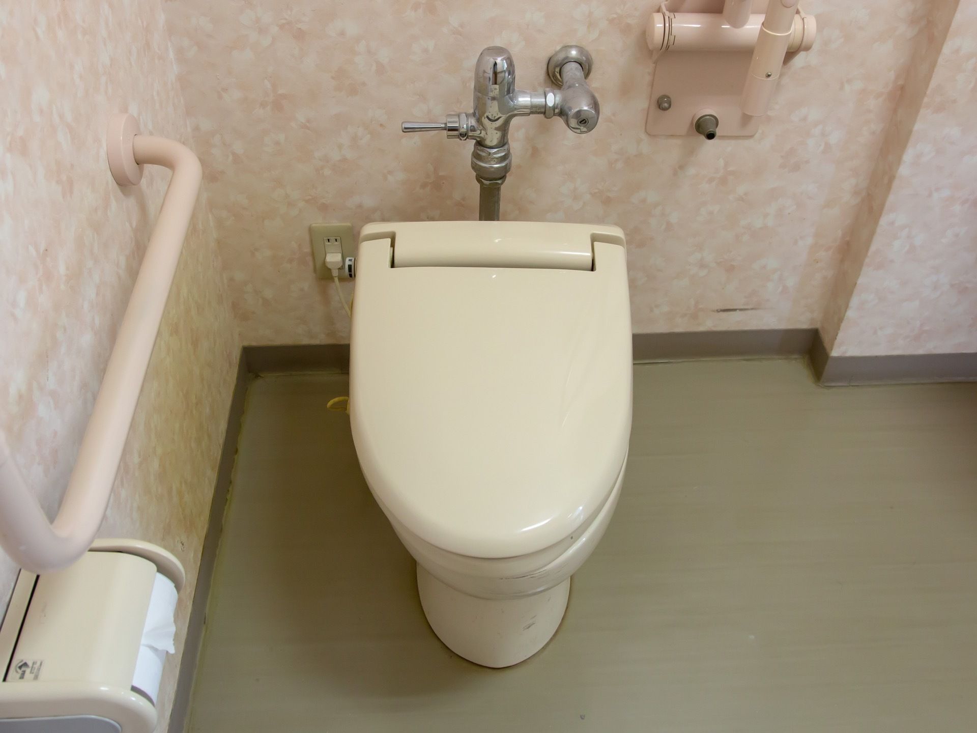 ピンク系の壁とモスグリーン床が落ち着いた雰囲気のトイレ。ゆとりあるスペースがあり、壁には手すりが取り付けられている。