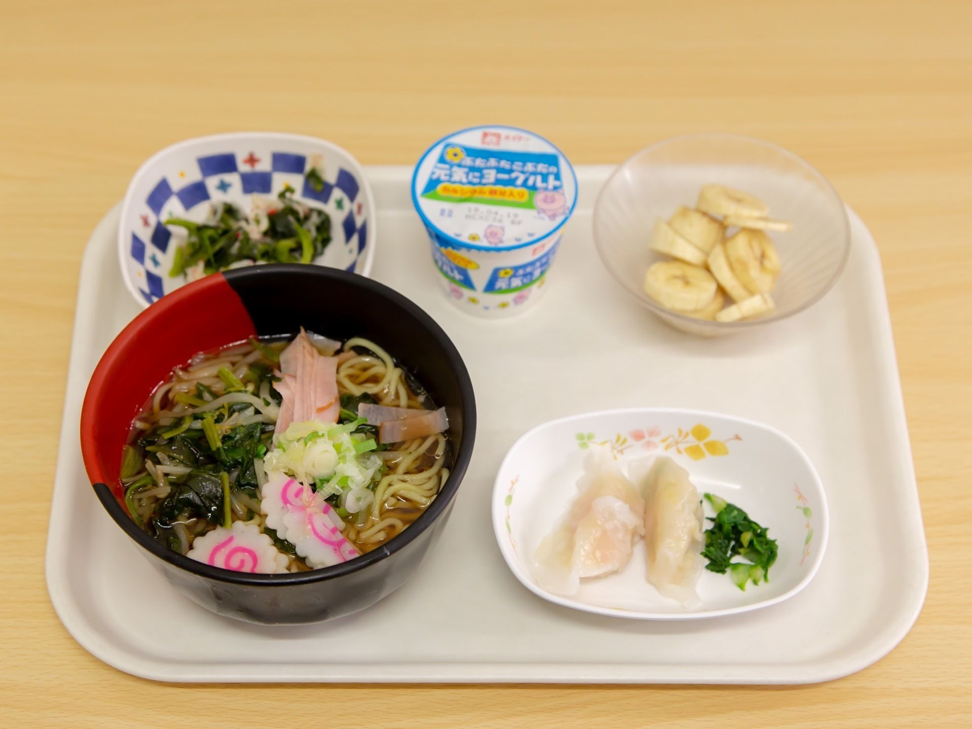 トレーの上に青菜やハム、ナルトがのったラーメンの他、水餃子、デザートのバナナやヨーグルト等、バランスのとれたメニューがのっている。
