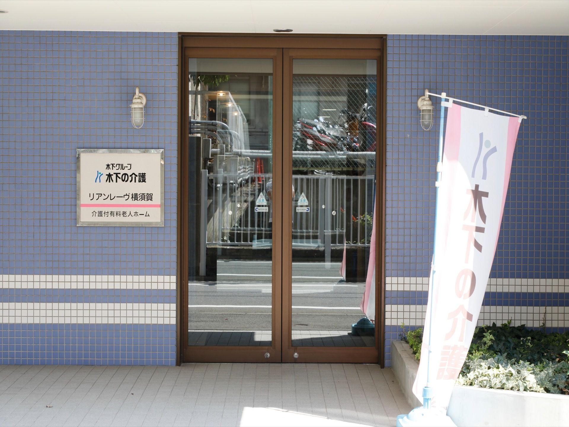 タイル貼りの外壁に大きな施設名が書かれている看板が取り付けられている。右側には花壇があり植物が植えてある。