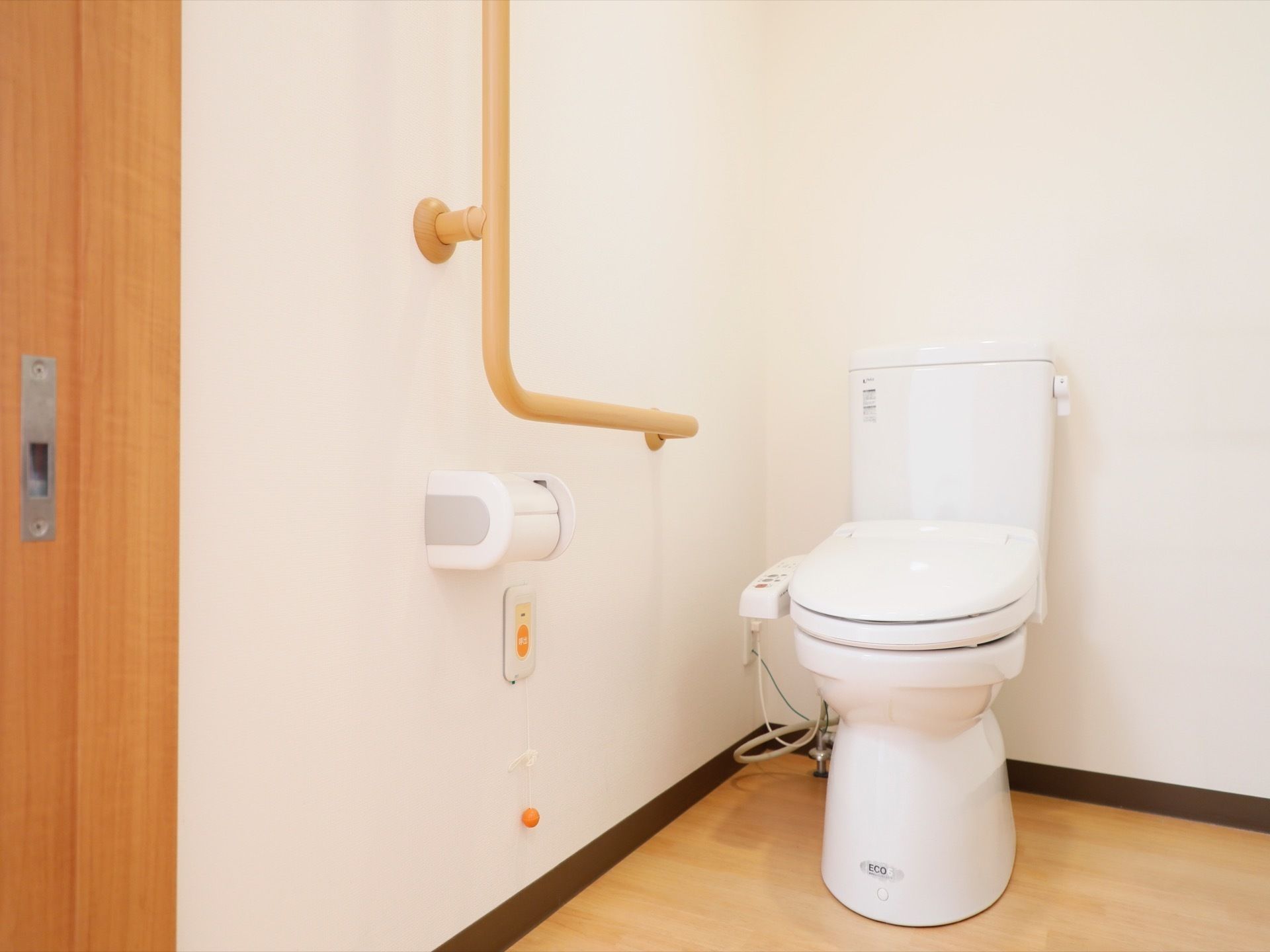 シンプルな便座にペーパーホルダーが１つ付いた広々したトイレ。温水洗浄ボタンは便器横に付いて、緊急ボタンも完備している。