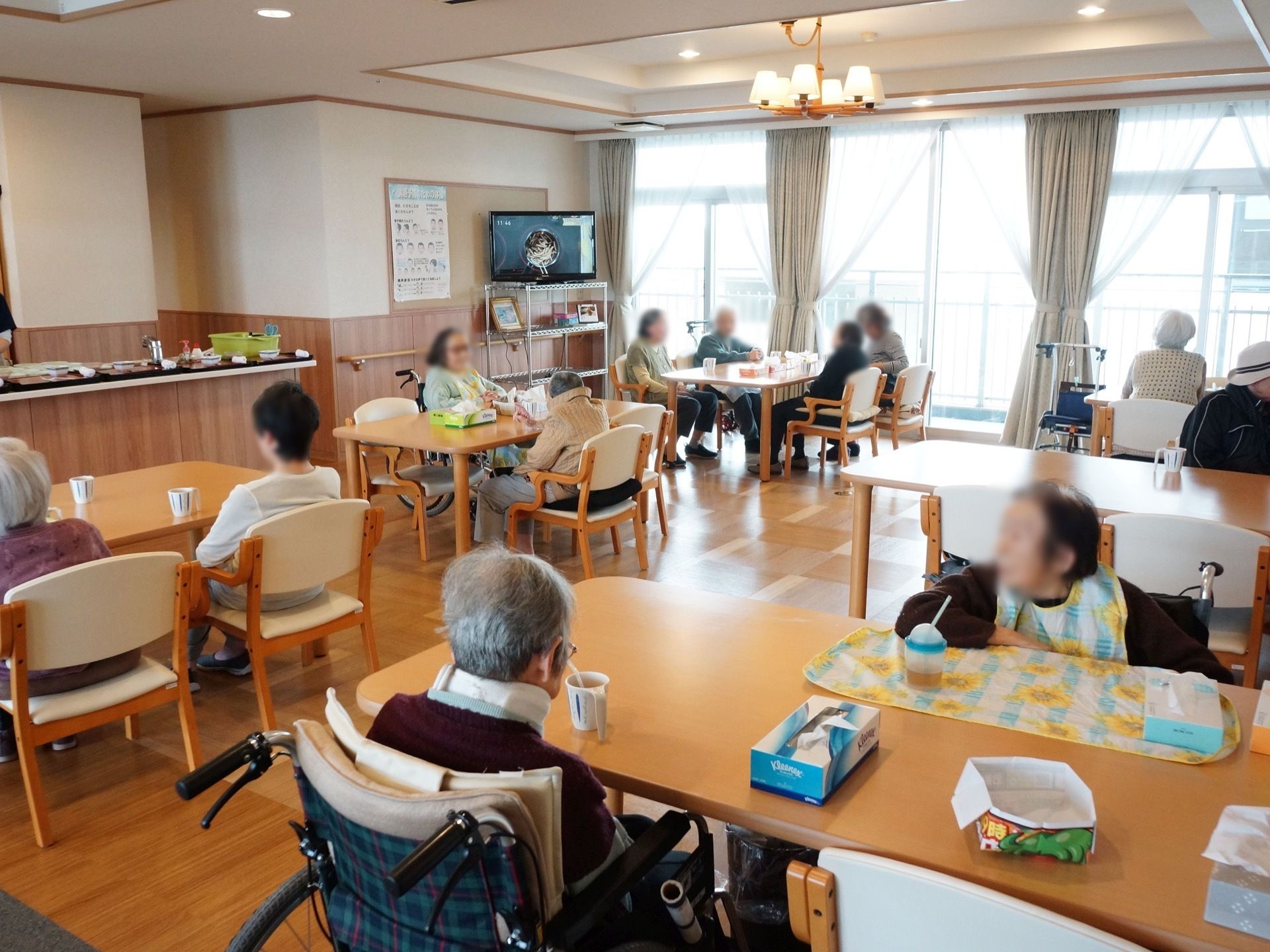 大きな窓がある明るいリビング。複数のテーブルや椅子が設置され、たくさんの入居者様がテレビを見たり、歓談したり、お茶を飲んだりしている。