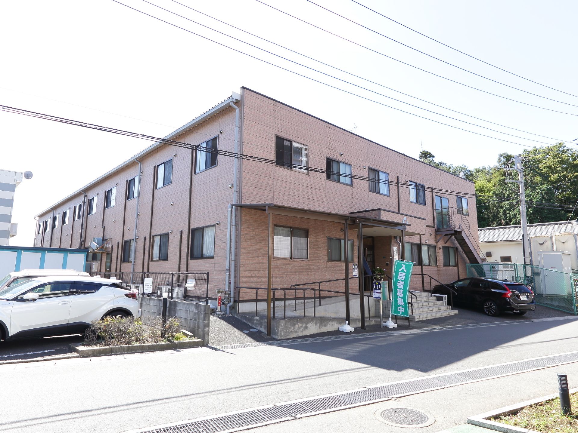 2階建ての建物で、玄関前にスロープと階段が設置されている。施設前に駐車場がある。外壁茶色である。入居者様募集ののぼりが掲げてある。