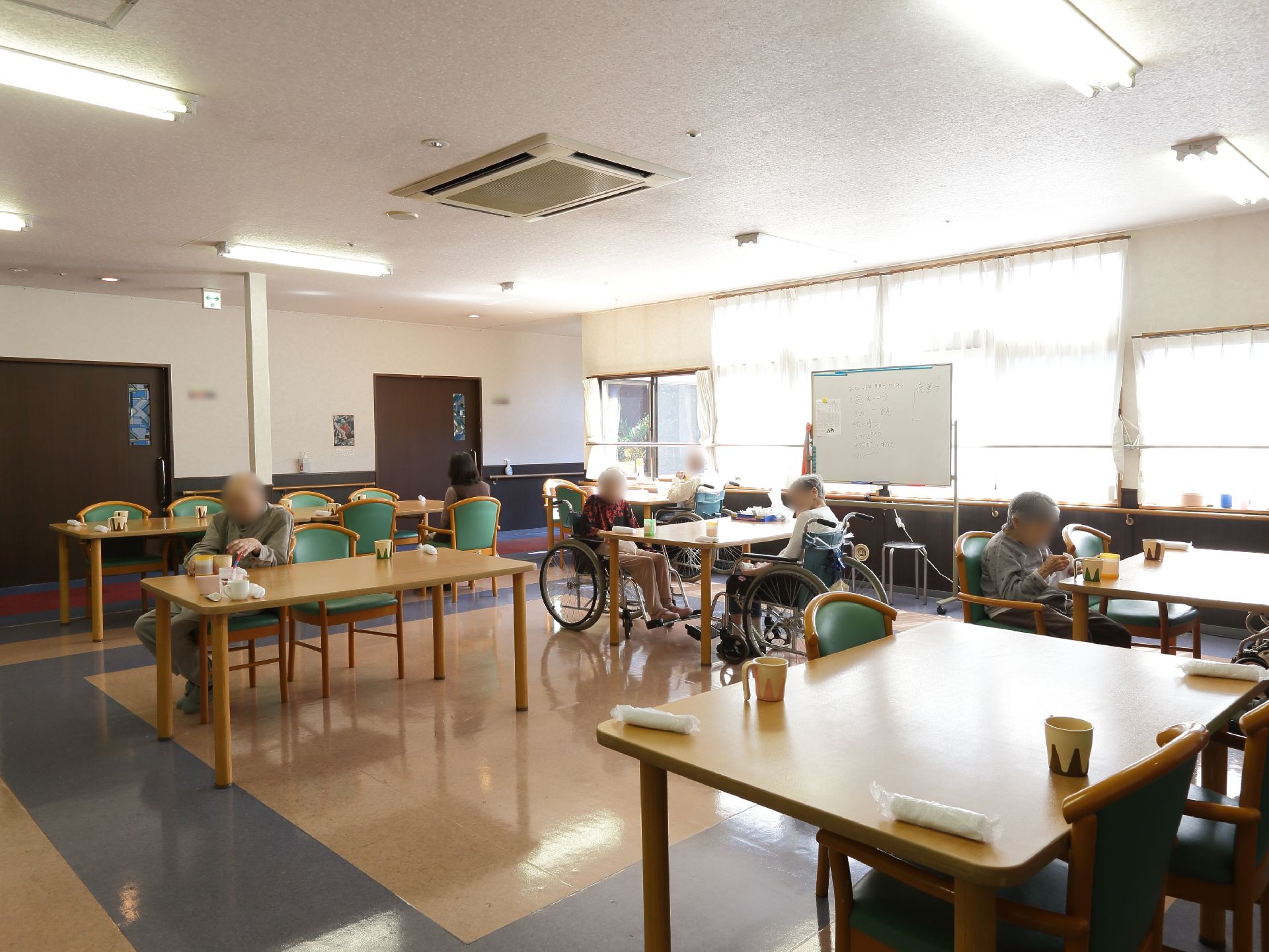 壁一面に窓があり明るい室内。木製のテーブル、椅子が幾つも置かれている。天井にはエアコンが設置されている。