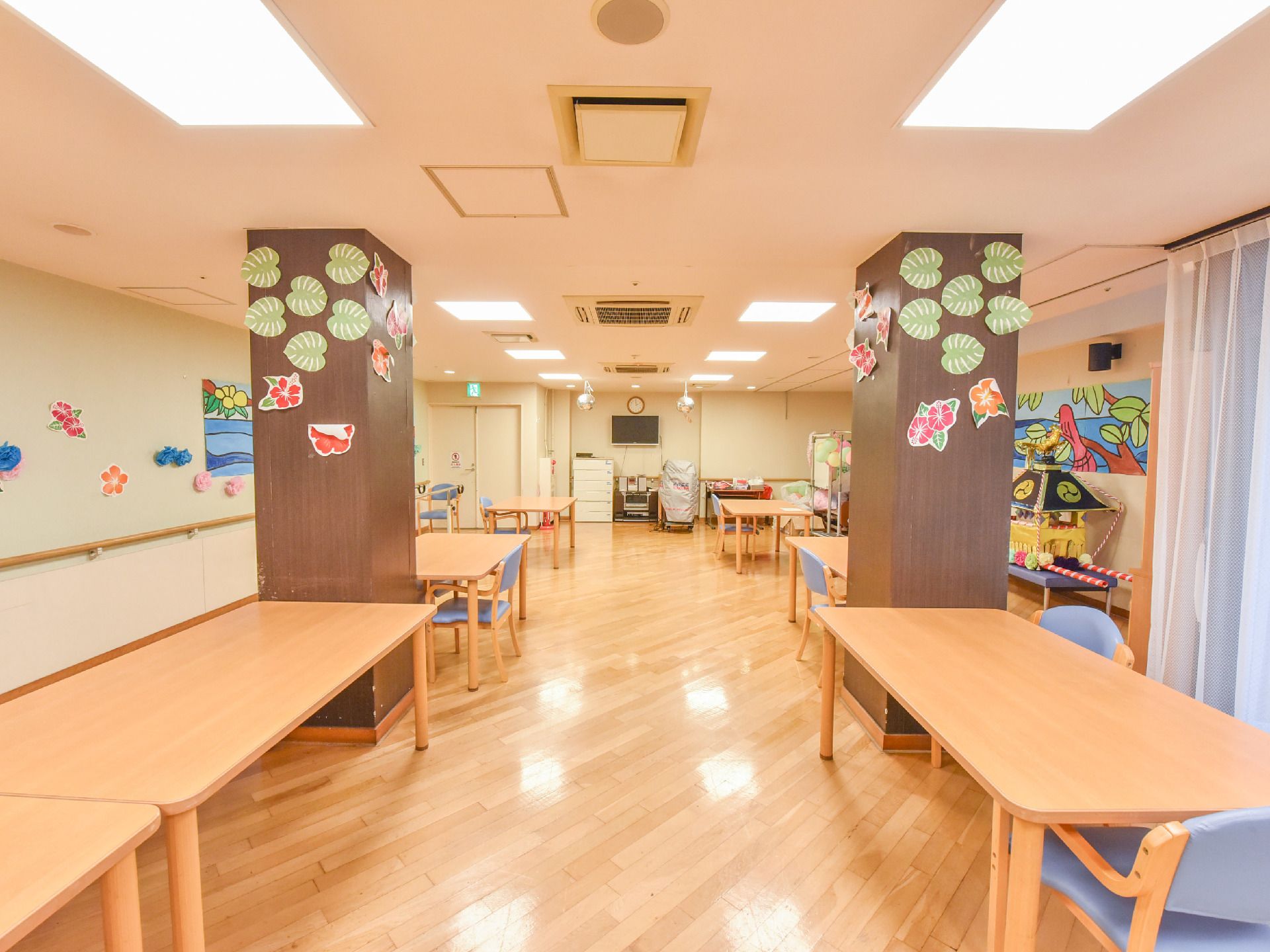 食堂の中央付近には、茶色の柱が2本あり、ピンクやオレンジ色の花が描かれた紙が飾り付けされている。奥にはテレビや時計がある。