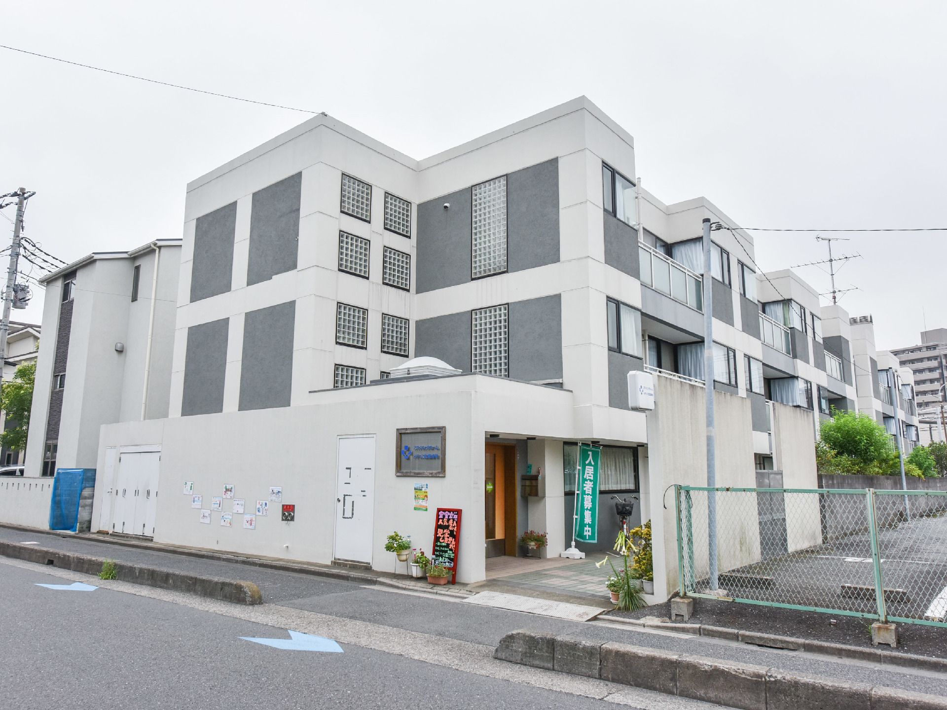 グレーと白の3階建ての建物。玄関は道路沿いではない。スタッフが手書きした看板を設置。他には、鉢植えもある。