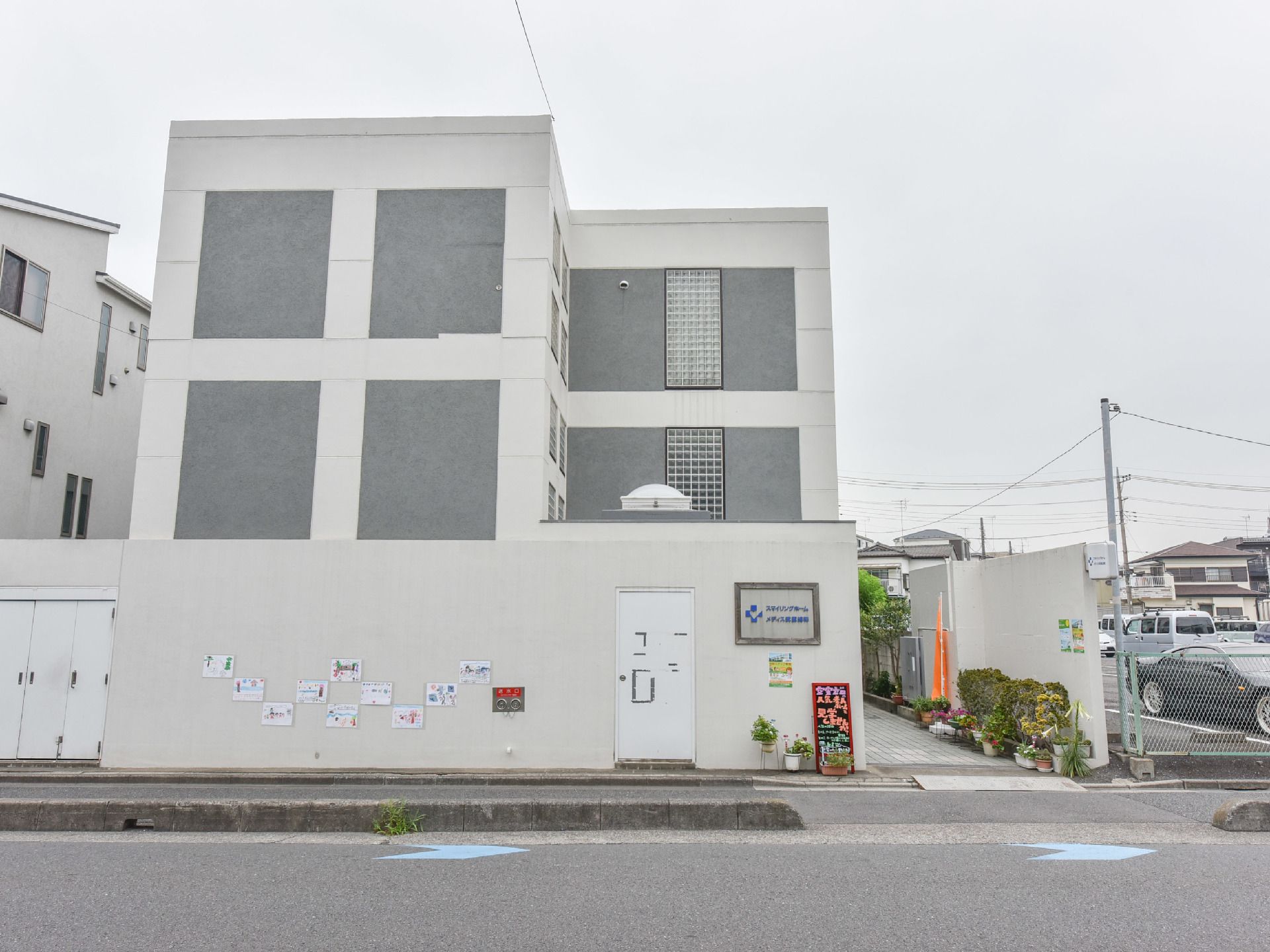 施設の外観はホワイト系の色とグレーのシンプルな建物になっている。建物は外壁で囲まれている。建物の前には歩道がある。